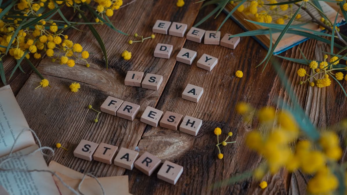 Wooden tiles spell 'Every day is a fresh start' surrounded by yellow flowers on rustic table.